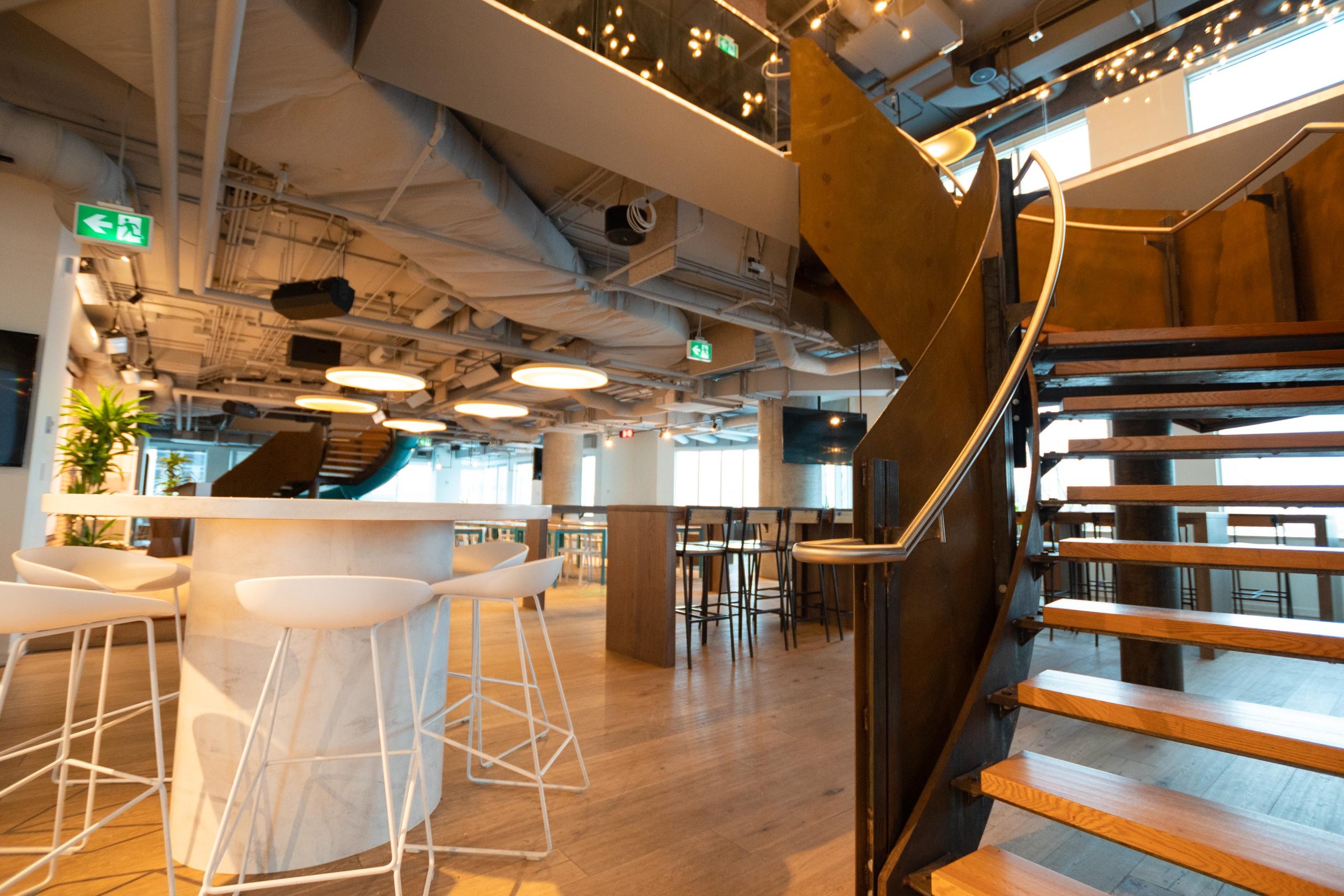 Modern co-working space with high tables, stools, and a spiral staircase.