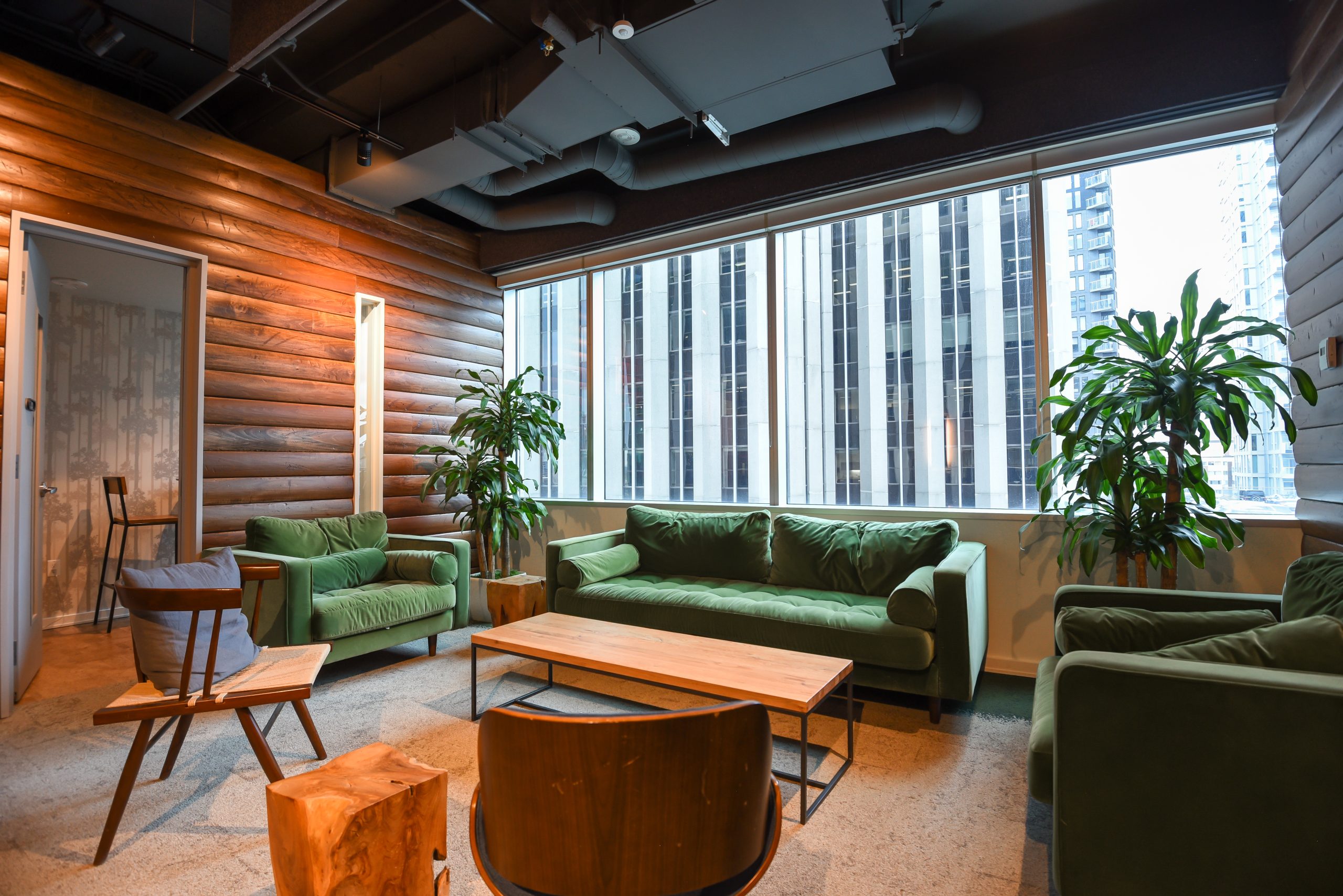 A cozy lounge area with green velvet sofas, wooden walls, and large windows overlooking a city.