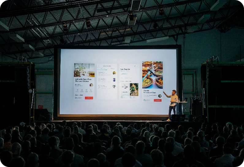 A speaker presenting on a large screen to a seated audience in a large, dark room.