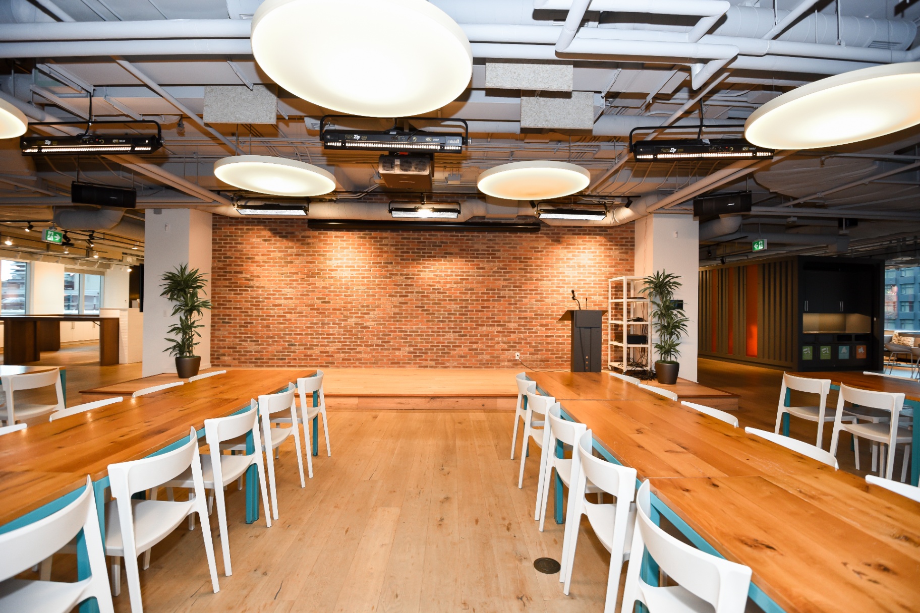 A spacious, contemporary conference room with long wooden tables, white chairs, a brick accent wall with a raised platform and podium.