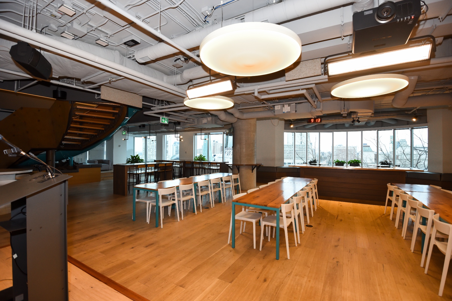 A spacious, brightly lit event space with long wooden tables, white chairs, a podium, and a staircase in the background.