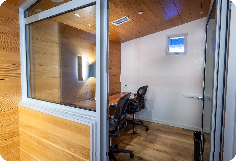 A brightly lit, sleek meeting room with long wooden tables, white chairs, and a brick accent wall with a podium.