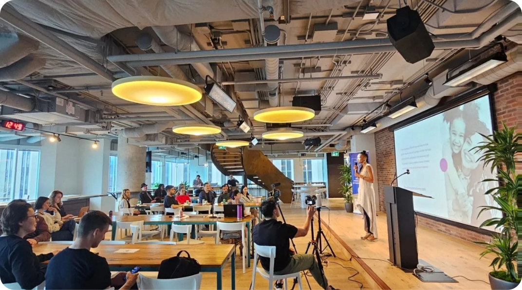 A woman presenting to an audience in a modern, industrial-style room with a projector screen.