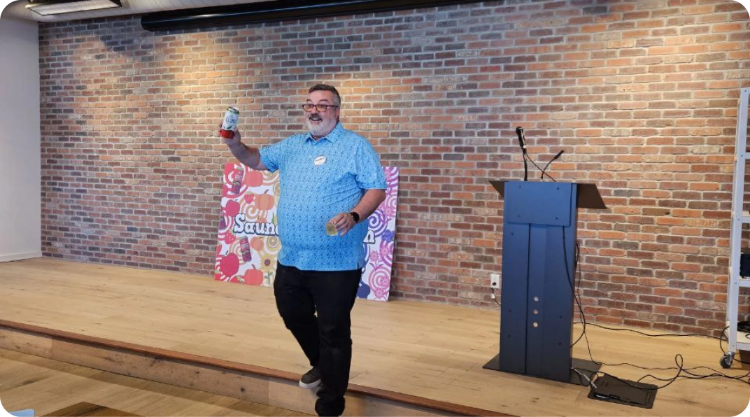 Man presenting on a stage with a brick wall backdrop, holding up a can.