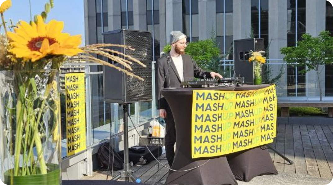 DJ performing on a rooftop with city views and black-covered tables with MASHUP banner. A networking event by TCC Canada