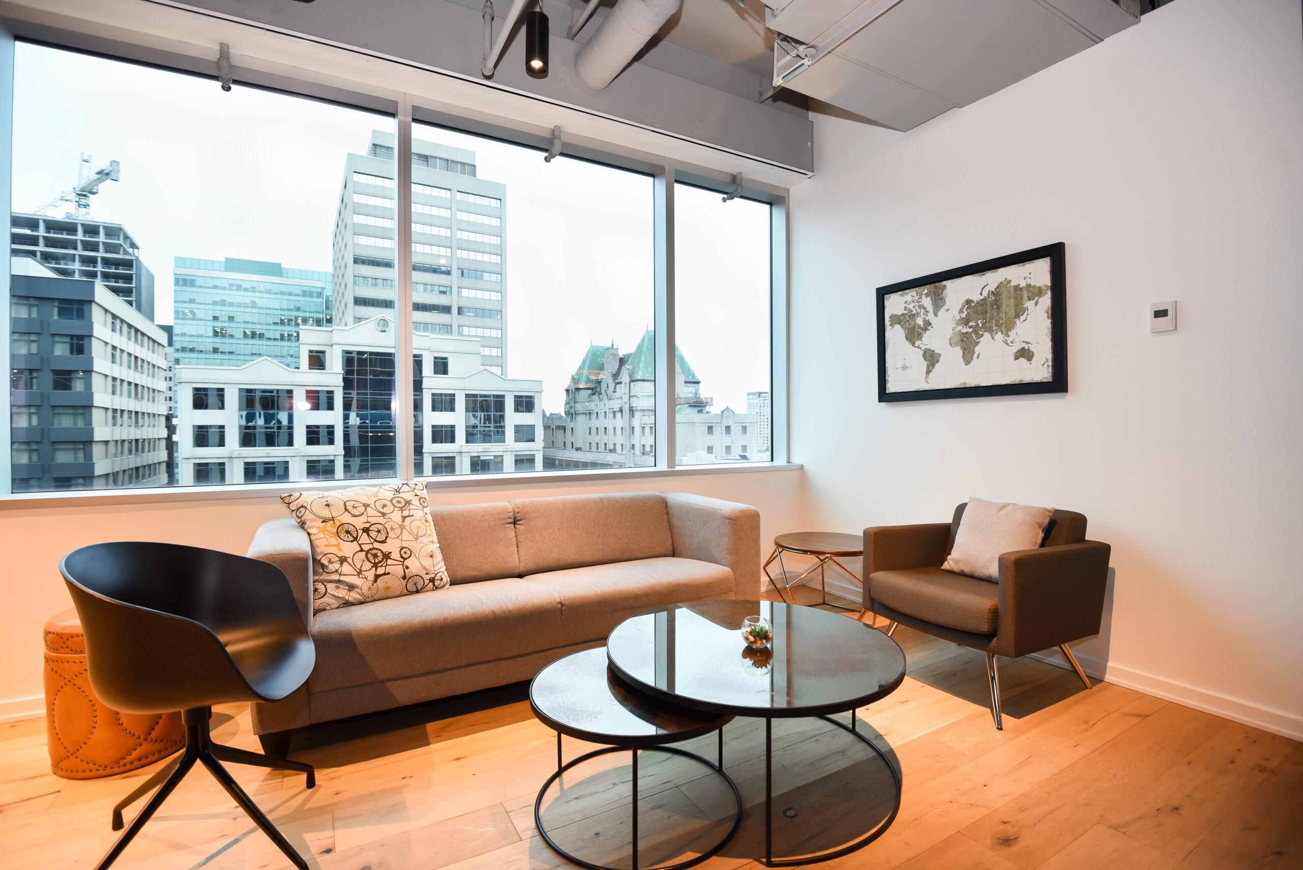A small lounge area with a beige sofa, a brown armchair, black tables, and a city view.