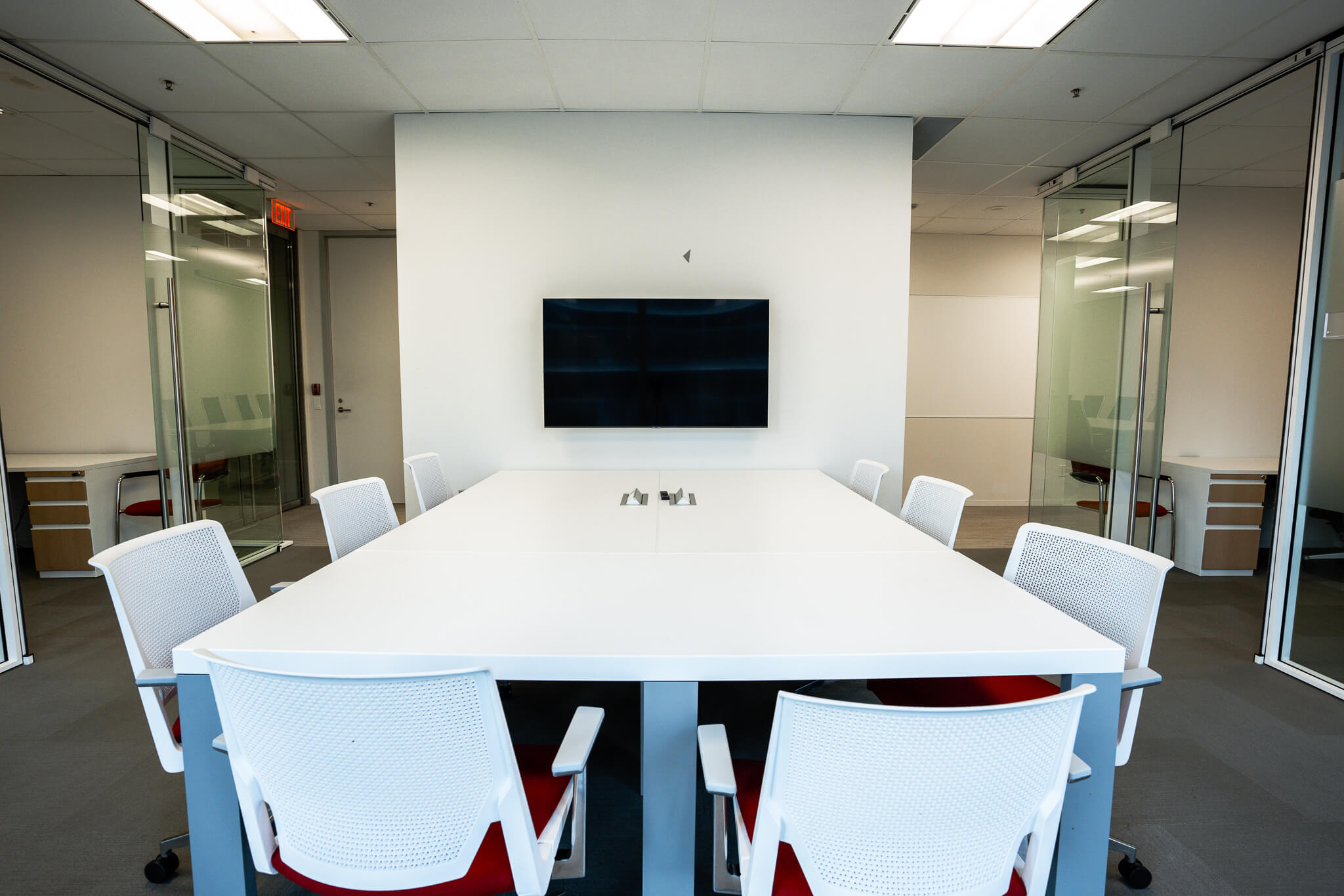 Modern, brightly lit conference room with a large white table, white chairs with red cushions, and a wall-mounted television.