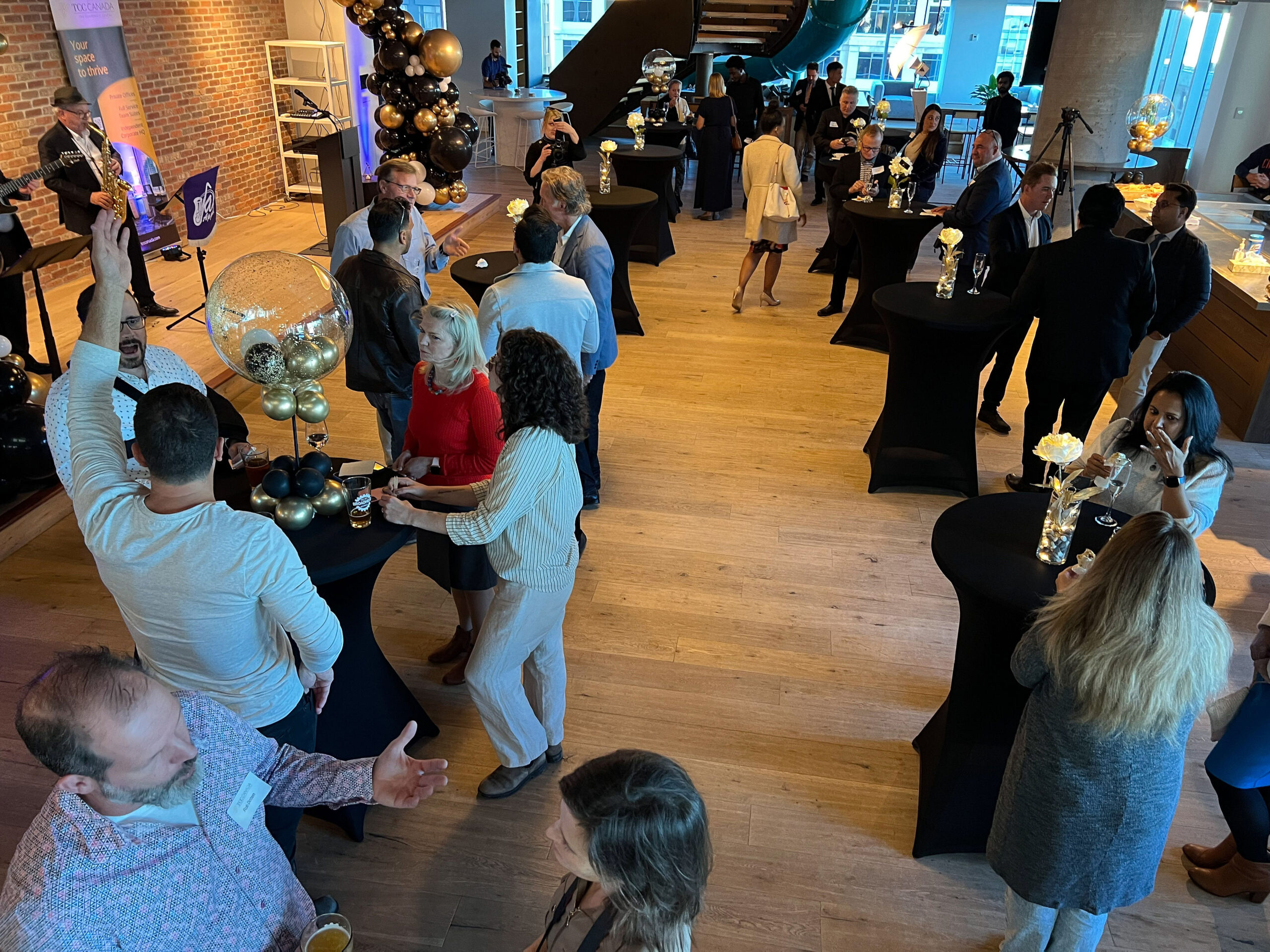 A crowded indoor event with people mingling around small black tables, balloons, and a musician performing.