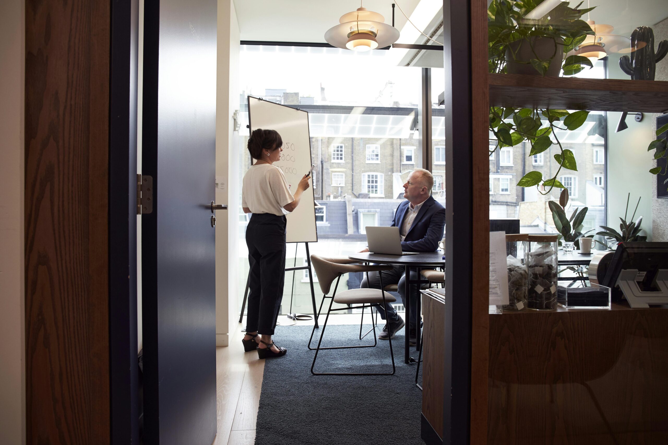 A woman appears to be giving a presentation on a whiteboard to a man with a laptop in a bright office.
