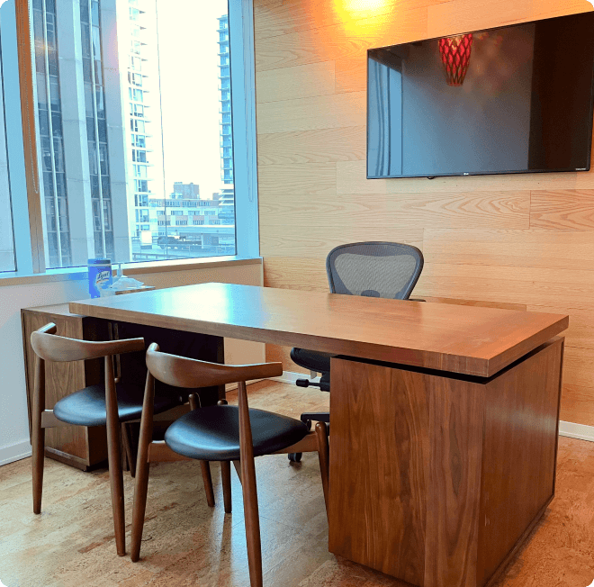 A stylish meeting room featuring a wooden conference table, four black chairs, and a basketball hoop decoration on the wooden wall. The room has a cityscape view through large windows.