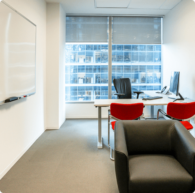 A cozy meeting area with a whiteboard, a desk, two red lounge chairs, and a black office chair. The office has a bright city view.