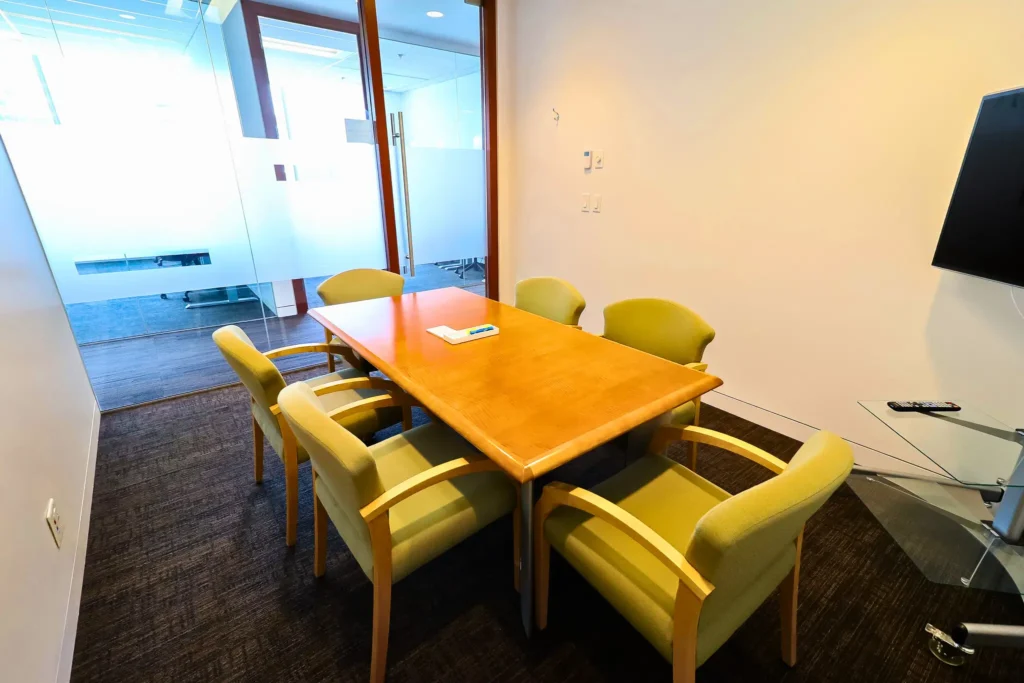 A small meeting room in The City Centre with a wooden table, six light green chairs, and a glass wall.